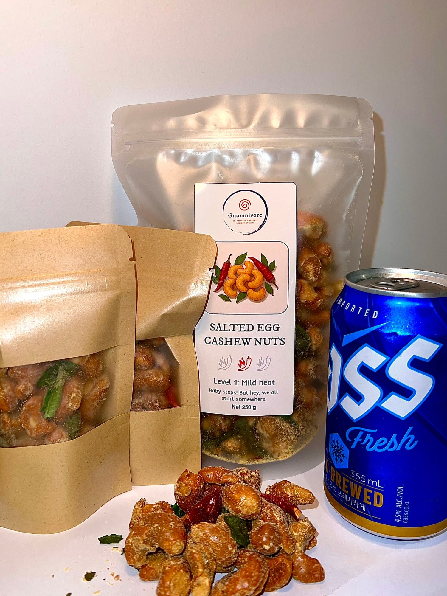 Assorted snacks including a bag of salted egg cashew nuts, a can of SGS Fresh, and a package of dried fruits on a white surface.